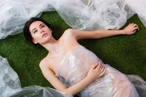 Top view of brunette woman wrapped in polyethylene lying on grass, ecology concept — Stock Photo