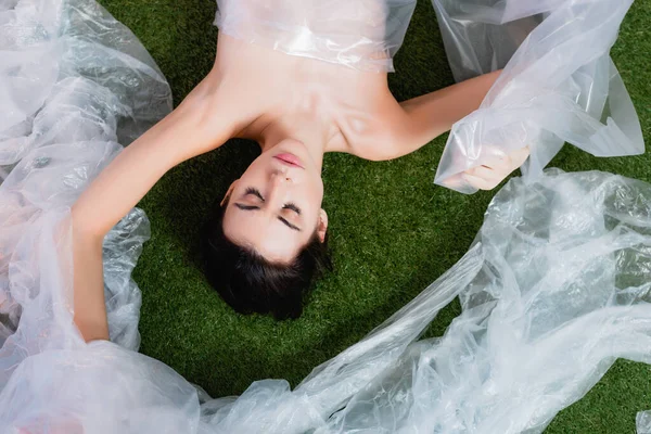 Top view of young brunette woman wrapped in polyethylene lying on grass, ecology concept — Stock Photo