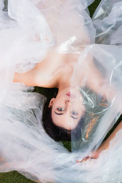 Top view of young brunette model wrapped in polyethylene lying and looking at camera, ecology concept — Stock Photo