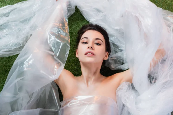 Top view of brunette model wrapped in polyethylene lying and looking at camera, ecology concept — Stock Photo