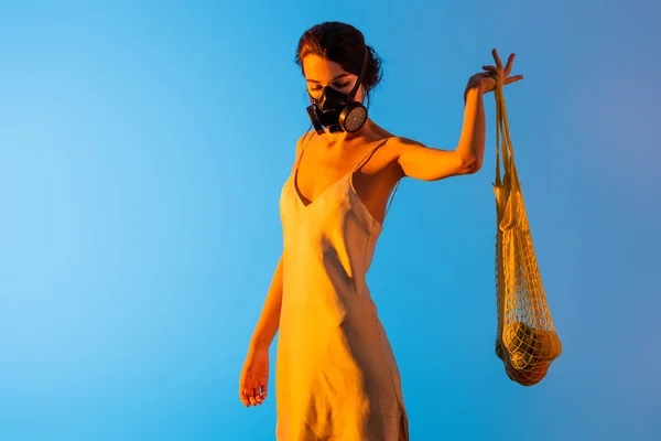 Jeune femme brune en masque à gaz et robe en soie tenant sac à ficelle réutilisable avec des fruits sur bleu — Photo de stock
