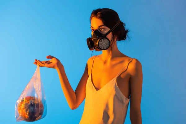 Femme en masque à gaz tenant sac en plastique avec globe sur bleu, concept écologie — Photo de stock