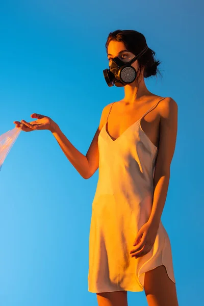 Жінка в газовій масці тримає пластиковий пакет на синьому, екологічна концепція — стокове фото