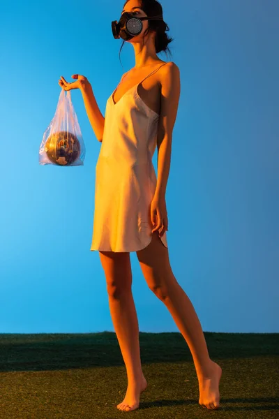 Femme pieds nus dans un masque à gaz tenant sac en plastique avec globe sur bleu, concept d'écologie — Photo de stock
