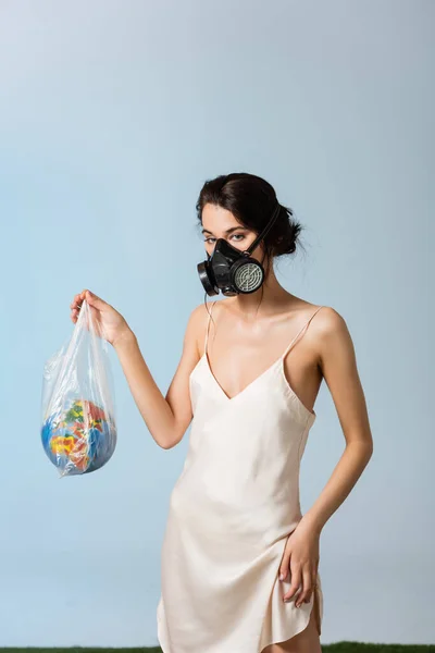 Женщина в противогазе, держащая пластиковый пакет с глобусом на сером, экологическая концепция — стоковое фото