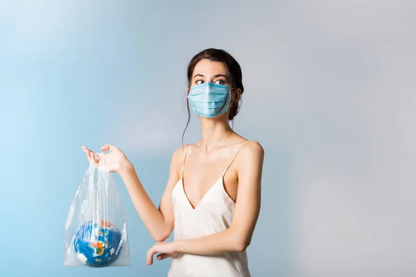 Modell in medizinischer Maske hält Plastiktüte mit Globus und schaut weg auf blaues, ökologisches Konzept — Stockfoto