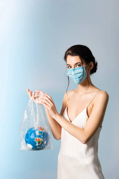 Mulher em máscara médica segurando saco de plástico com globo e olhando para a câmera em azul, conceito de ecologia — Fotografia de Stock