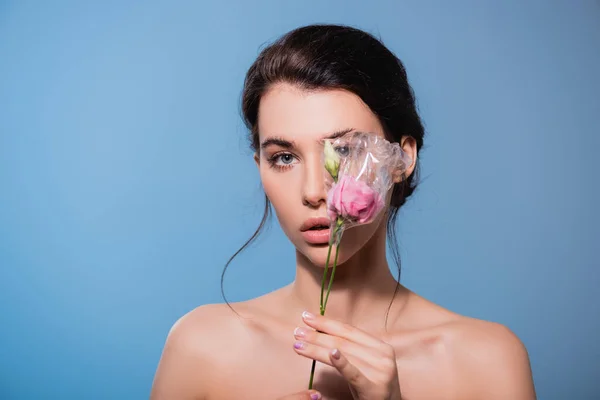 Young and naked woman covering eye with flowers in polyethylene isolated on blue, ecology concept — Stock Photo