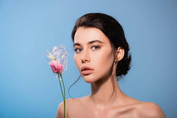 Young and naked woman holding flowers in polyethylene isolated on blue, ecology concept — Stock Photo