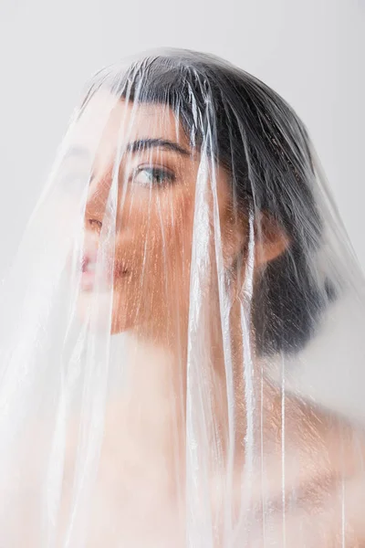 Brunette woman looking at camera through polyethylene isolated on grey, ecology concept — Stock Photo