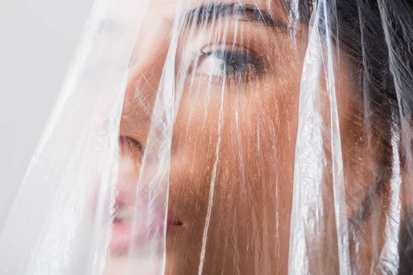 Nahaufnahme einer brünetten Frau, die durch Polyethylen in die Kamera blickt, isoliert auf grau, ökologisches Konzept — Stockfoto