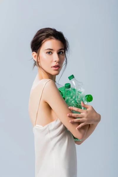 Брюнетка в шелковом платье с пустыми и пластиковыми бутылками, изолированными на сером, экологическая концепция — стоковое фото