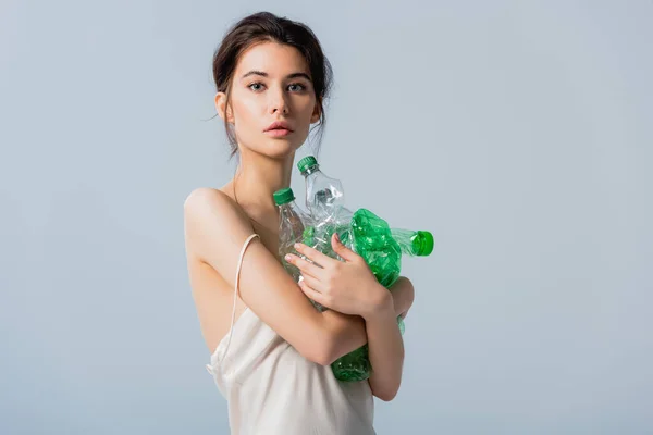 Morena mujer en vestido de seda mirando a la cámara mientras sostiene botellas de plástico aisladas en gris, concepto de ecología - foto de stock