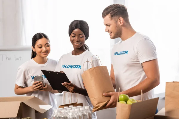 Selective focus of multiethnic volunteers looking at clipboard near packages with food and water in charity center — Stock Photo