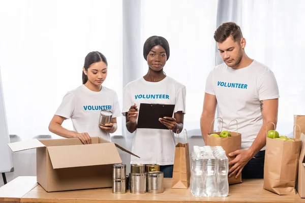 Multiethnic volunteers with clipboard standing near paper bags and boxes with food in charity center — Stock Photo