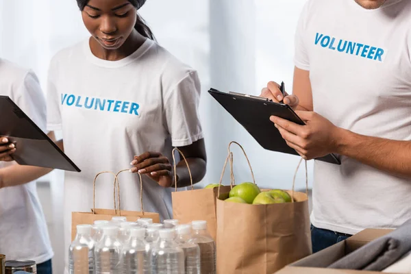 Enfoque selectivo de voluntarios multiétnicos con portapapeles cerca de paquetes con comida y botellas de agua - foto de stock