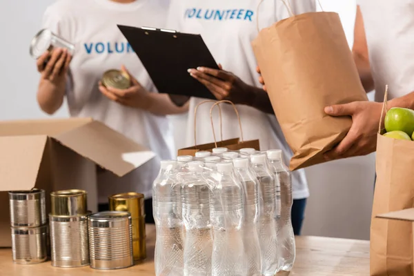 Vista recortada de voluntarios multiétnicos con portapapeles cerca de botellas de agua y paquetes con comida en el centro de caridad - foto de stock