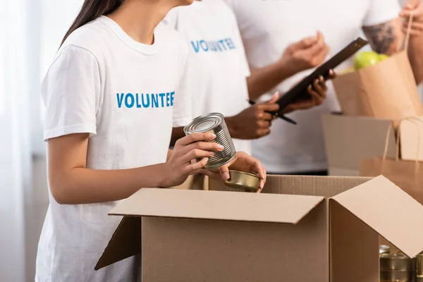 Vista recortada de una mujer sosteniendo latas de lata cerca de voluntarios multiétnicos con portapapeles en el centro de caridad - foto de stock