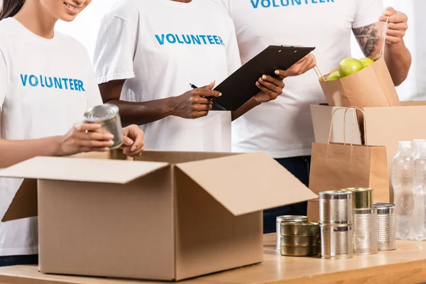 Cropped view of multicultural volunteers with clipboard packing food on table in charity center — Stock Photo