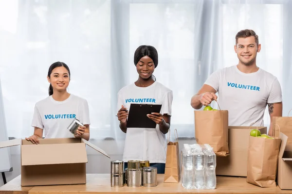 Multiethnic volunteers with clipboard and food looking at camera near packages in charity center — Stock Photo