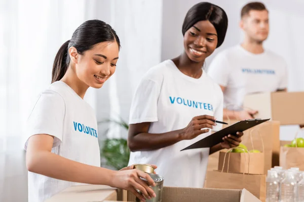 Selective focus of asian volunteer putting tin cans in box near african american woman with clipboard in charity center — Stock Photo