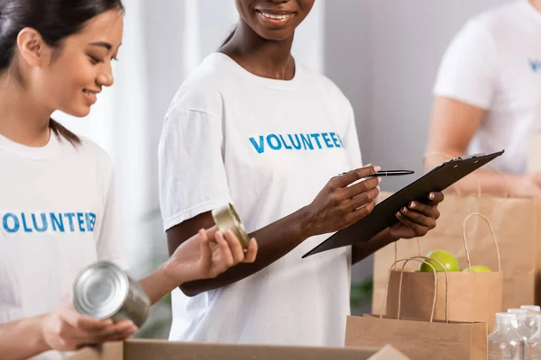 Enfoque selectivo del voluntario afroamericano sosteniendo la pluma y el portapapeles cerca de la mujer asiática con latas y paquetes en el centro de caridad - foto de stock