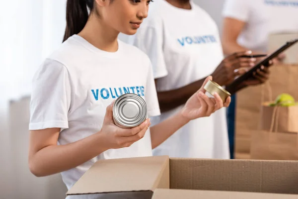 Selective focus of asian volunteer holding tin cans near cardboard box in charity center — Stock Photo