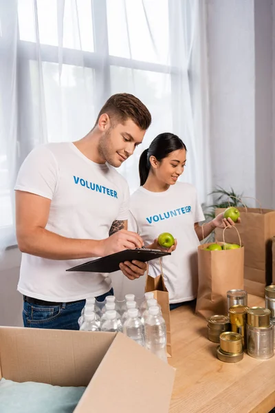 Selective focus of multiethnic volunteers writing on clipboard and holding apples near tin cans and bottles of water on table — Stock Photo