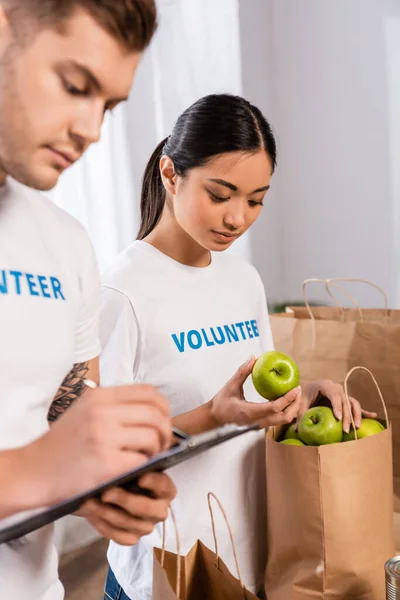 Enfoque selectivo de asiático voluntario celebración de manzanas cerca de hombre escribir en portapapeles en centro de caridad - foto de stock