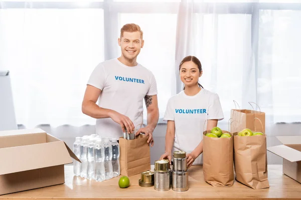 Мультиэтнические волонтеры смотрят в камеру возле еды, бутылки с водой и пакеты на столе в благотворительном центре — стоковое фото