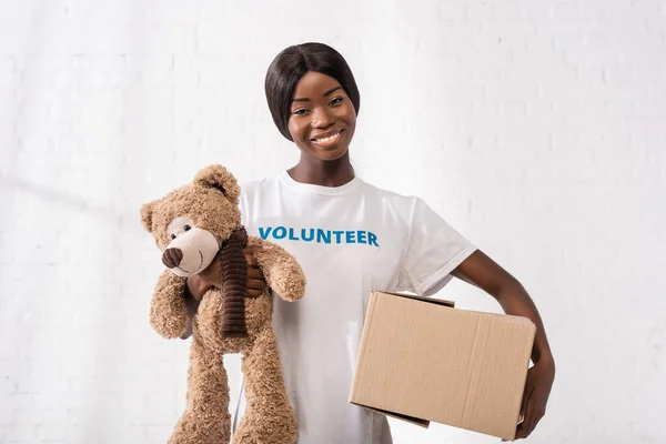 Volontaire afro-américain regardant la caméra tout en tenant boîte en carton et jouet doux — Photo de stock