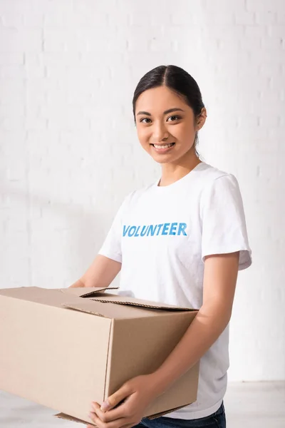 Voluntario asiático mirando la cámara mientras sostiene la caja de cartón - foto de stock