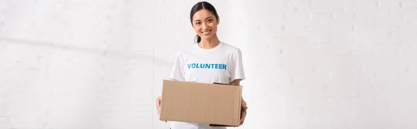 Panoramic crop of asian woman with lettering volunteer on t-shirt holding package — Stock Photo