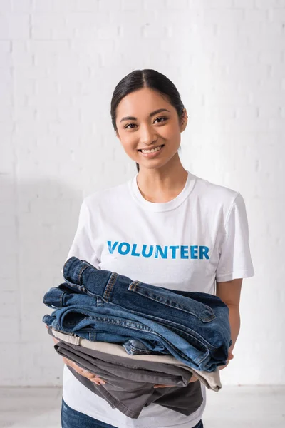 Asian volunteer holding clothes and looking at camera in charity center — Stock Photo