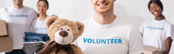 Панорамний знімок людини, що тримає м'яку іграшку біля багатоетнічних добровольців у благодійному центрі — стокове фото