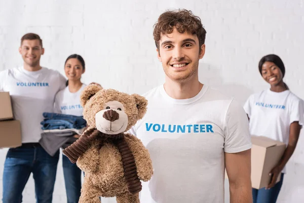Foco seletivo do homem com letras voluntárias na camiseta segurando brinquedo macio no centro de caridade — Fotografia de Stock