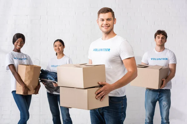 Selective focus of man holding carton boxes near volunteers with paper bag and clothes at background — Stock Photo