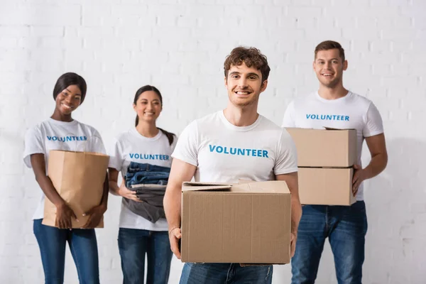 Enfoque selectivo del hombre con paquete de pie cerca de voluntarios multiétnicos en el centro de caridad - foto de stock