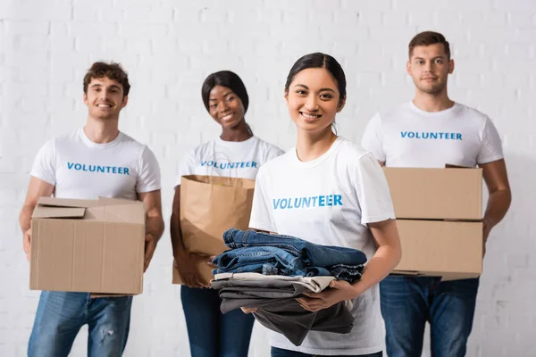 Selective focus of asian woman holding clothes near multicultural volunteers in charity center — Stock Photo