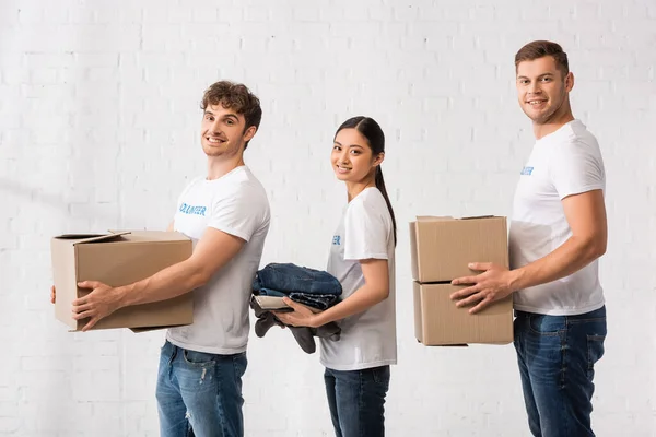 Multiethnic volunteers holding carton boxes and clothes while looking at camera in charity center — Stock Photo