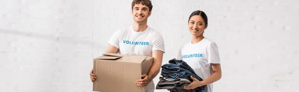 Imagen horizontal de voluntarios multiculturales sosteniendo caja de cartón y ropa en el centro de caridad - foto de stock