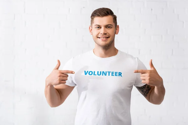 Young volunteer pointing at lettering on white t-shirt and looking at camera — Stock Photo