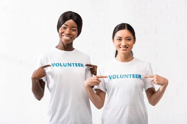 African american and asian volunteers pointing with fingers at t-shirts — Stock Photo