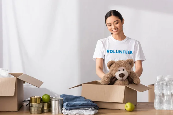 Asian volunteer putting soft toy in carton package near clothes and tin cans in charity center — Stock Photo