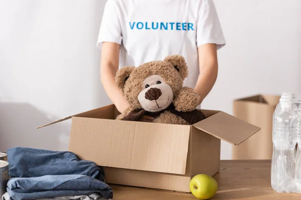 Cropped view of young volunteer putting soft toy in cardboard box near clothes, apple and bottles of water in charity center — Stock Photo