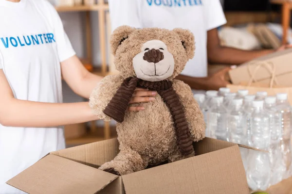Vista recortada del voluntario poniendo juguete suave en caja de cartón cerca de paquetes con botellas de agua en el centro de caridad - foto de stock