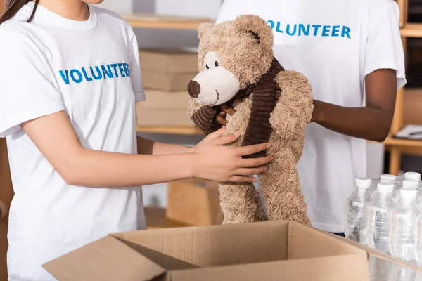 Cropped view of multicultural volunteers holding soft toy near carton box and bottles of water in charity center — Stock Photo