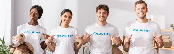 Cultivo horizontal de jóvenes voluntarios multiétnicos que señalan con los dedos las letras en las camisetas y muestran los pulgares hacia arriba en el centro de caridad - foto de stock