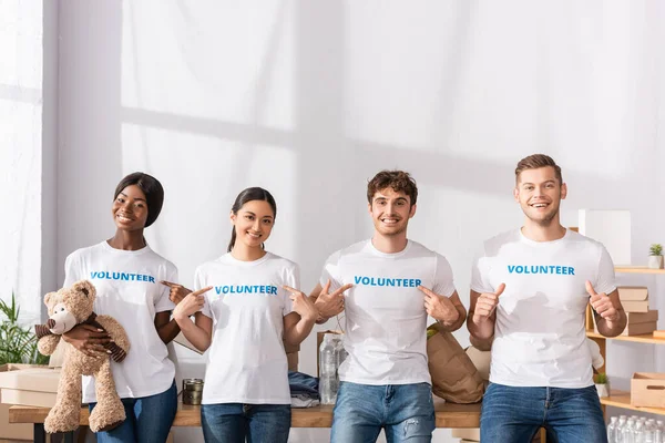 Multiethnic volunteers pointing with fingers at t-shirts and showing like in charity center — Stock Photo