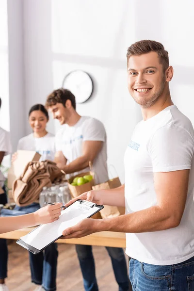 Selektiver Fokus von Freiwilligen, die in der Nähe einer Frau in die Kamera schauen, die auf Klemmbrett im Charity Center schreibt — Stockfoto